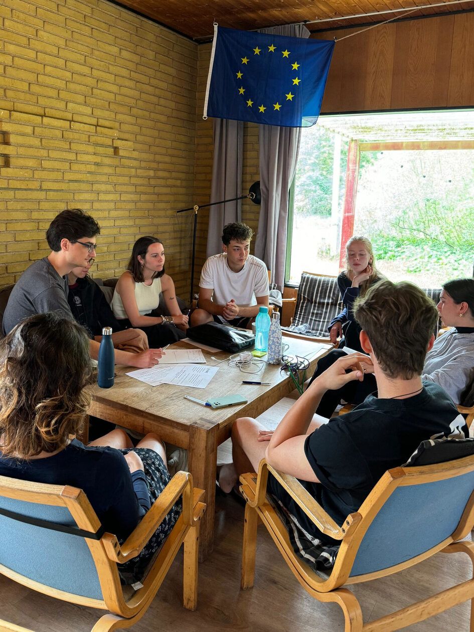 Sieben Jugendliche sitzen um einen Tisch, über ihnen hängt eine Europa-Fahne.