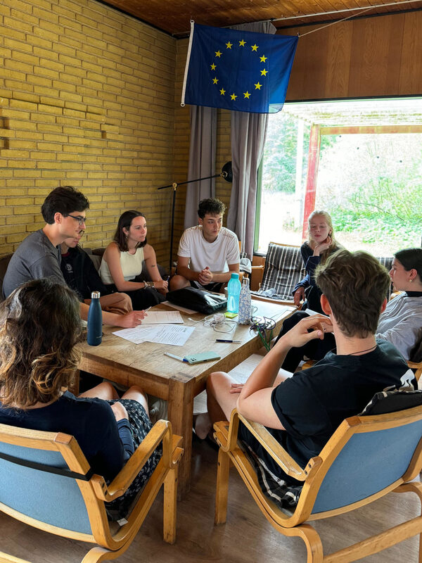 Sieben Jugendliche sitzen um einen Tisch, über ihnen hängt eine Europa-Fahne.