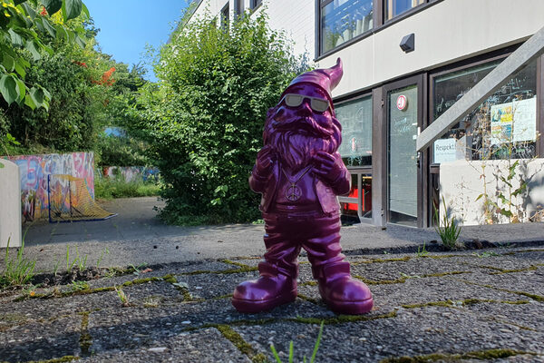 Ein Gartenzwerg steht auf einem Boden mit Betonplatten, im Hintergrund ist eine Glastür zu sehen.