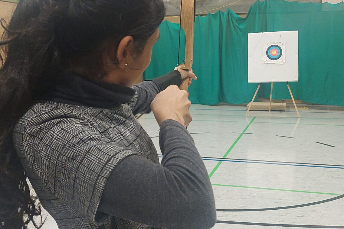 ein Mädchen zielt mit einem Bogen auf eine Scheibe.