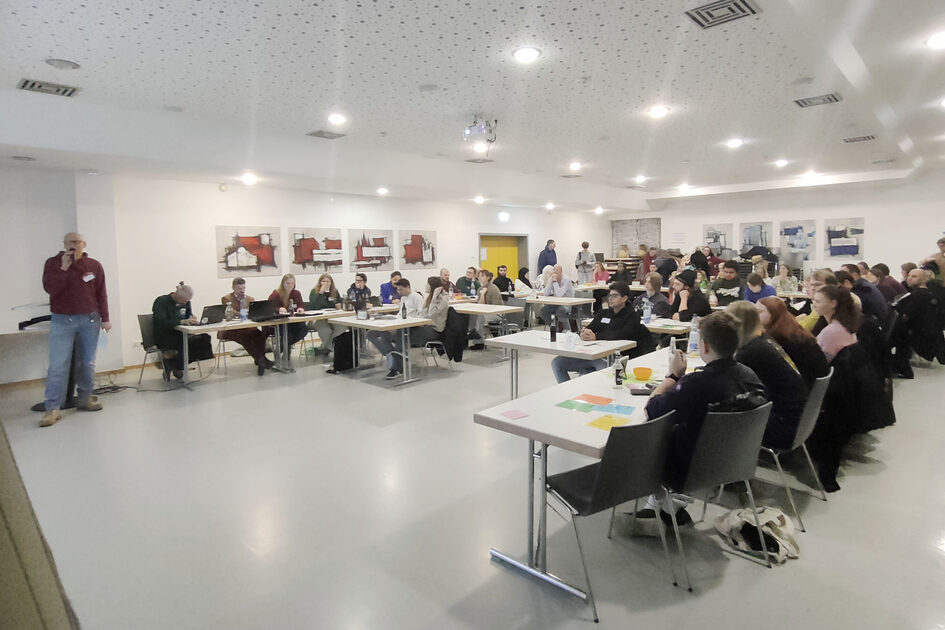 Viele junge Menschen sitzen an Tischen, ein Mann steht.