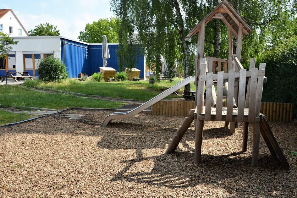 Das Außengelände der Kindertagesstätte. Im Vordergrund steht ein Kletterhaus mit einer Rutsche. Im Hintergrund ist das Gebäude