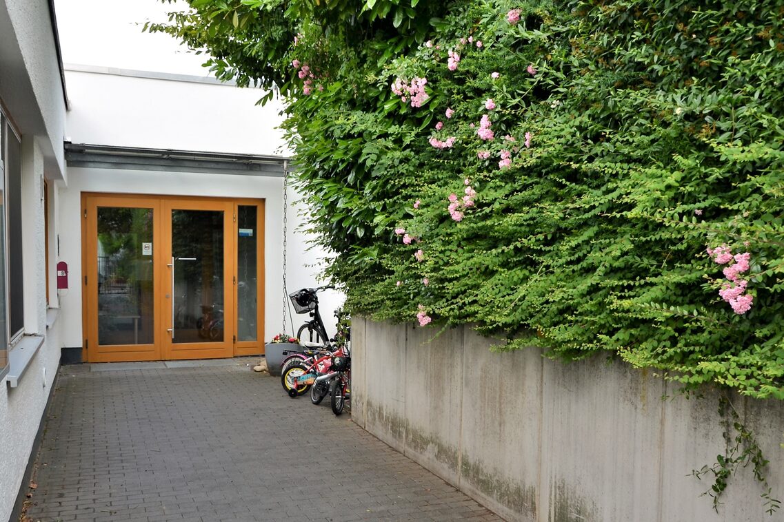 Der Eingangsbereich der Kindertagesstätte. Rechts wird der Zugang von einer MAuer mit einer großen Hecke begrenzt.