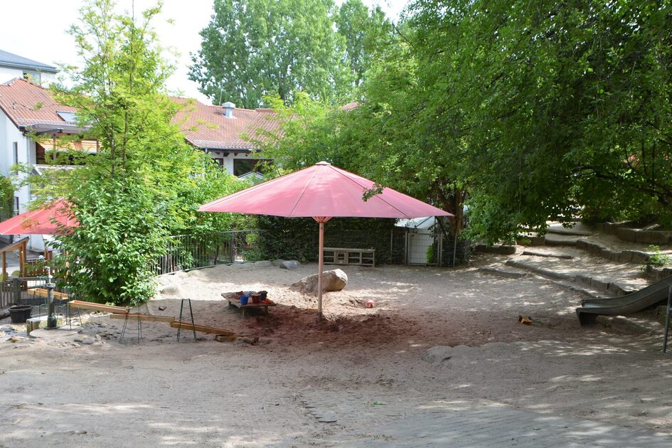 Das Außengelände der Kindertagesstätte mit dem Sandspielbereich und einem aufgespannten roten Sonnenschirm