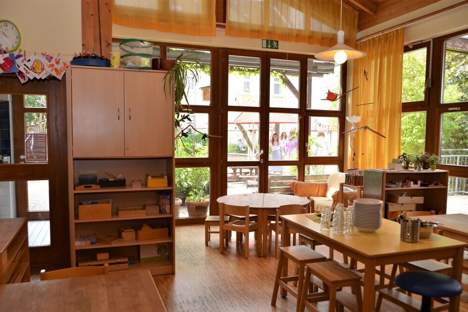 Der Gruppenraum im Elementarbereich mit Tischen und Stühlen, einem Regal und einer großen Fensterfront. Von der Decke hängt ein Mobile´