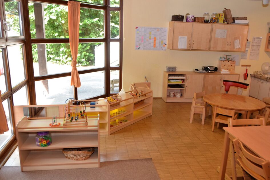 Ein Gruppenraum in der Krippe mit kleinen Tischen und Stühlen, Regalen und eine große Fensterfront