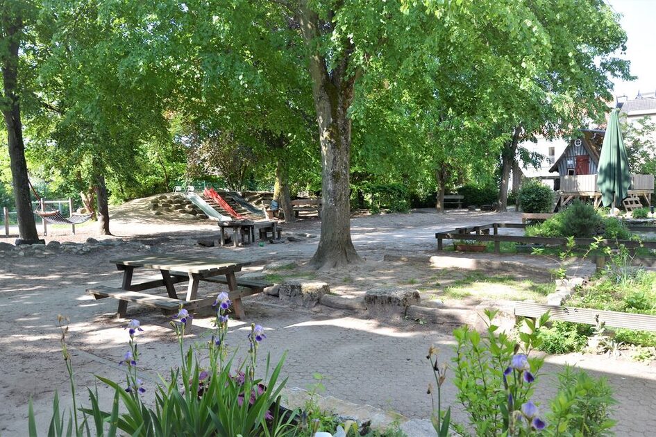 Das Außengelände der Kindertagesstätte. Im Bildmittelpunkt steht ein großer Baum mit einem Tisch und einer Bank. Im Hintergrund ist eine Rutsche