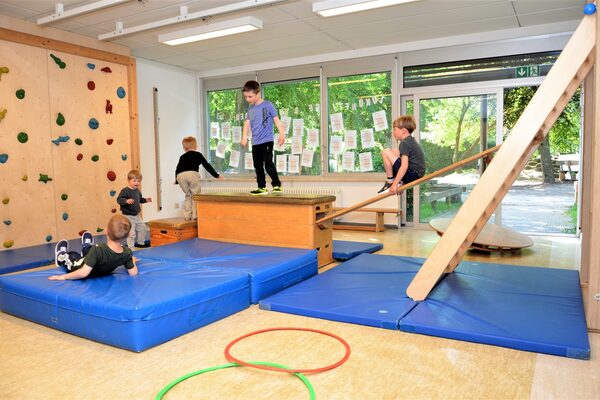 Der Bewegungsraum mit großen blauen Matten auf dem Boden auf denen Kinder turnen. An der wand ist eine Kletterwand