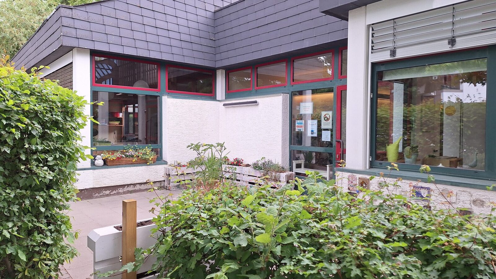 Der Eingangsbereich der Kindertagesstätte. Im Vordergrund eine Hecke und aufrecht stehende Paletten zum Bespflanzen