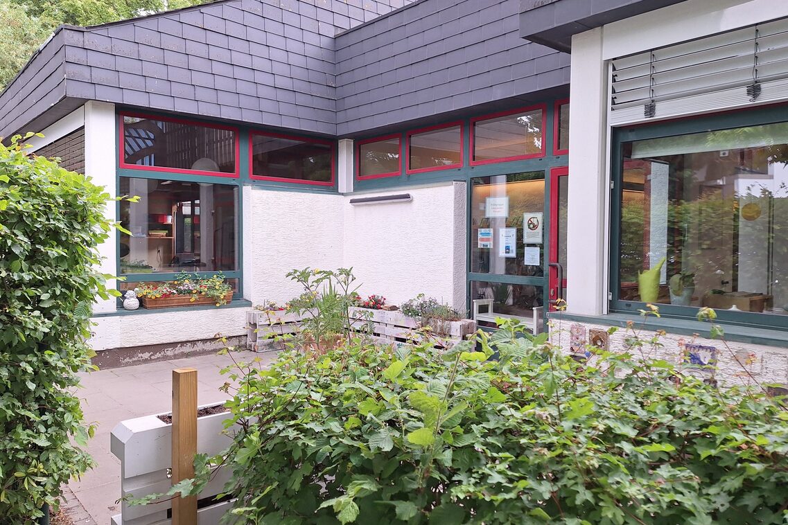 Der Eingangsbereich der Kindertagesstätte. Im Vordergrund eine Hecke und aufrecht stehende Paletten zum Bespflanzen