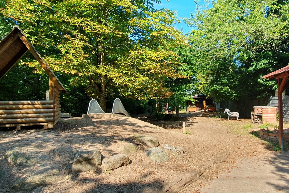 Das Außengelände mit einer Rutsche, die von einem Hügel hinab führt. Auf dem Hügel steht ein Spielhaus aus Holz. Im Hintergrund sind Bäume