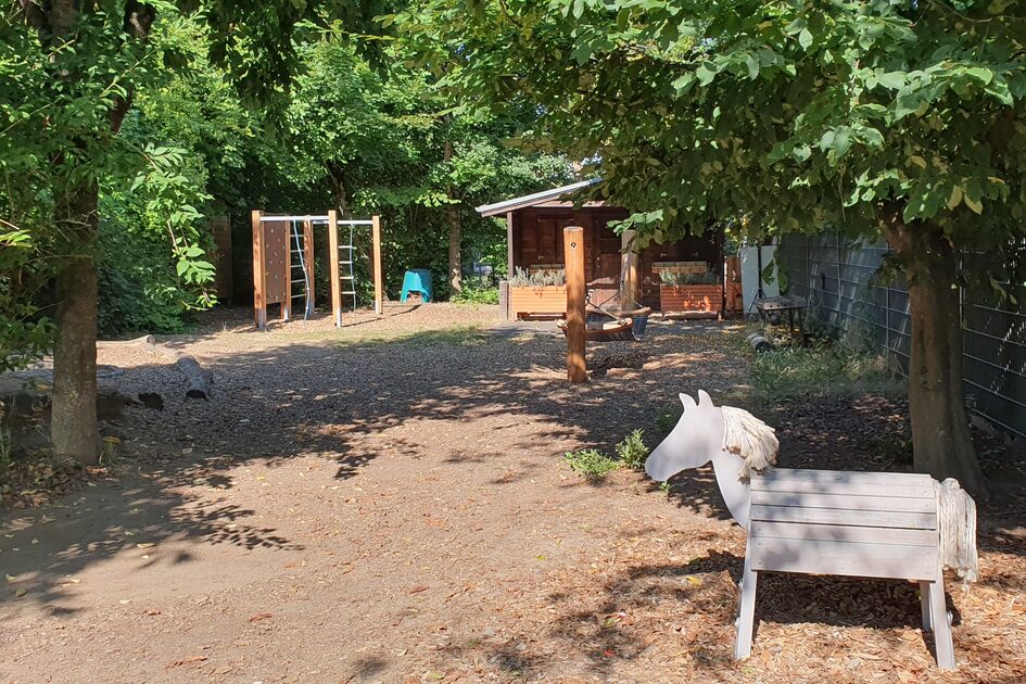 Der hintere Teil des Außengeländes mit einem Klettergerüst und einem Spielhaus. Im Vordergrund steht ein Holzpferd. Umrahmt wird das Gelände von Bäumen.