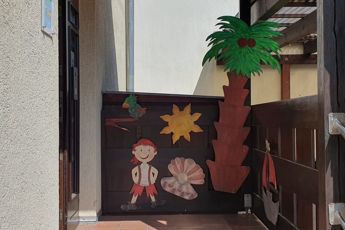 Der Eingang des Kinderhauses. Die Tür ist links und an der Holzumrandung ist eine Palme , ein Pirat und Seetiere.
