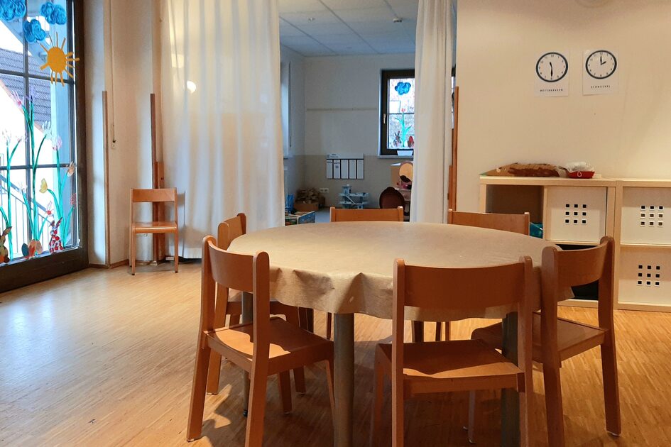 Der Gruppenraum des Kinderhauses. Im Vordergrund steht ein Tisch mit Stühlen. An der Wand im Hintergrund steht ein Regal. Links ist eine Fensterfront.