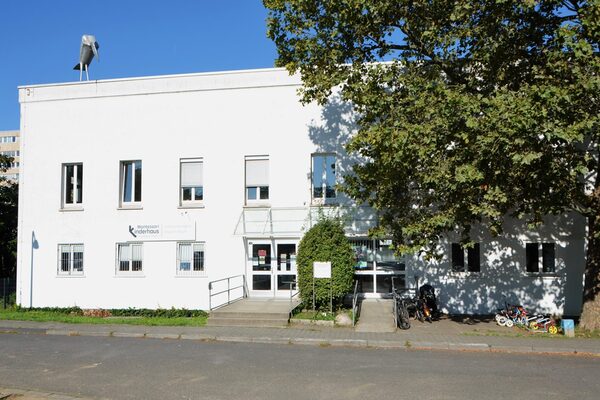 Das gebäude der Kindertagesstätte mit dem Eingang. Links steht ein hoher Baum und auf dem Dach steht ein Rabe aus Blech.