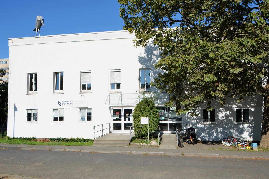 Das gebäude der Kindertagesstätte mit dem Eingang. Links steht ein hoher Baum und auf dem Dach steht ein Rabe aus Blech.