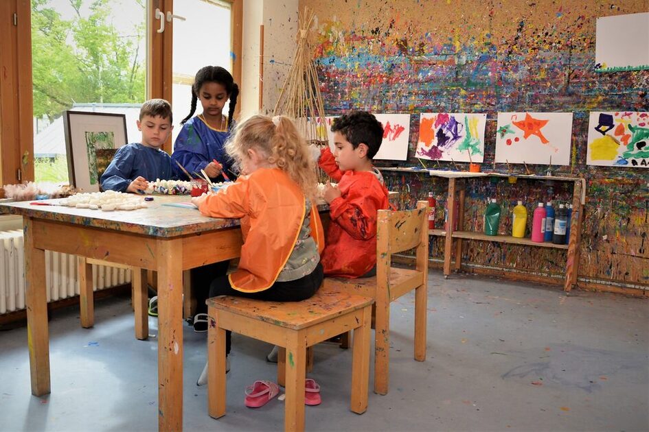 DAs Kunstatelier. Kinder sitzen an einem Tisch und malen. An der Wand hängen selbst gefertigte Kunstwerke