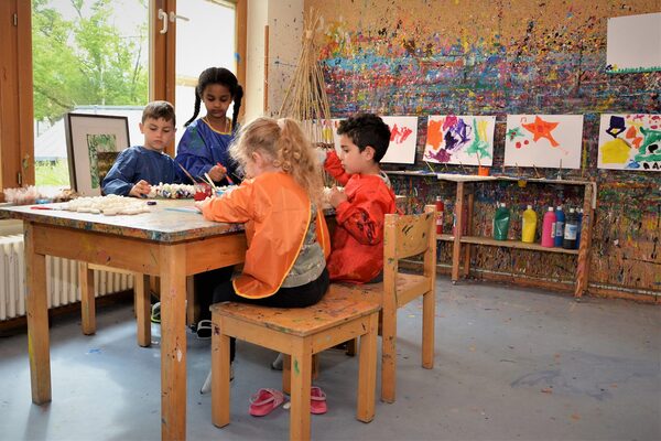 DAs Kunstatelier. Kinder sitzen an einem Tisch und malen. An der Wand hängen selbst gefertigte Kunstwerke