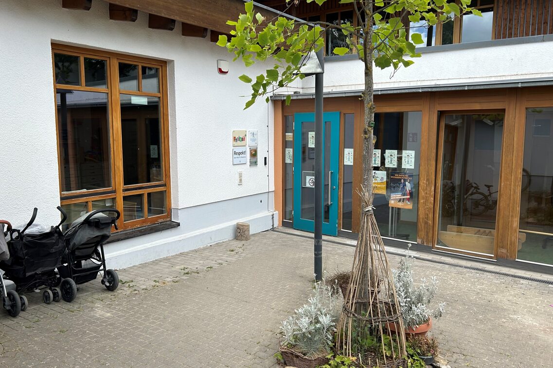 Der Eingang der Kindertagesstätte mit einem Baum im Vordergrund. Auf der linken Bildseite stehen Kinderwagen