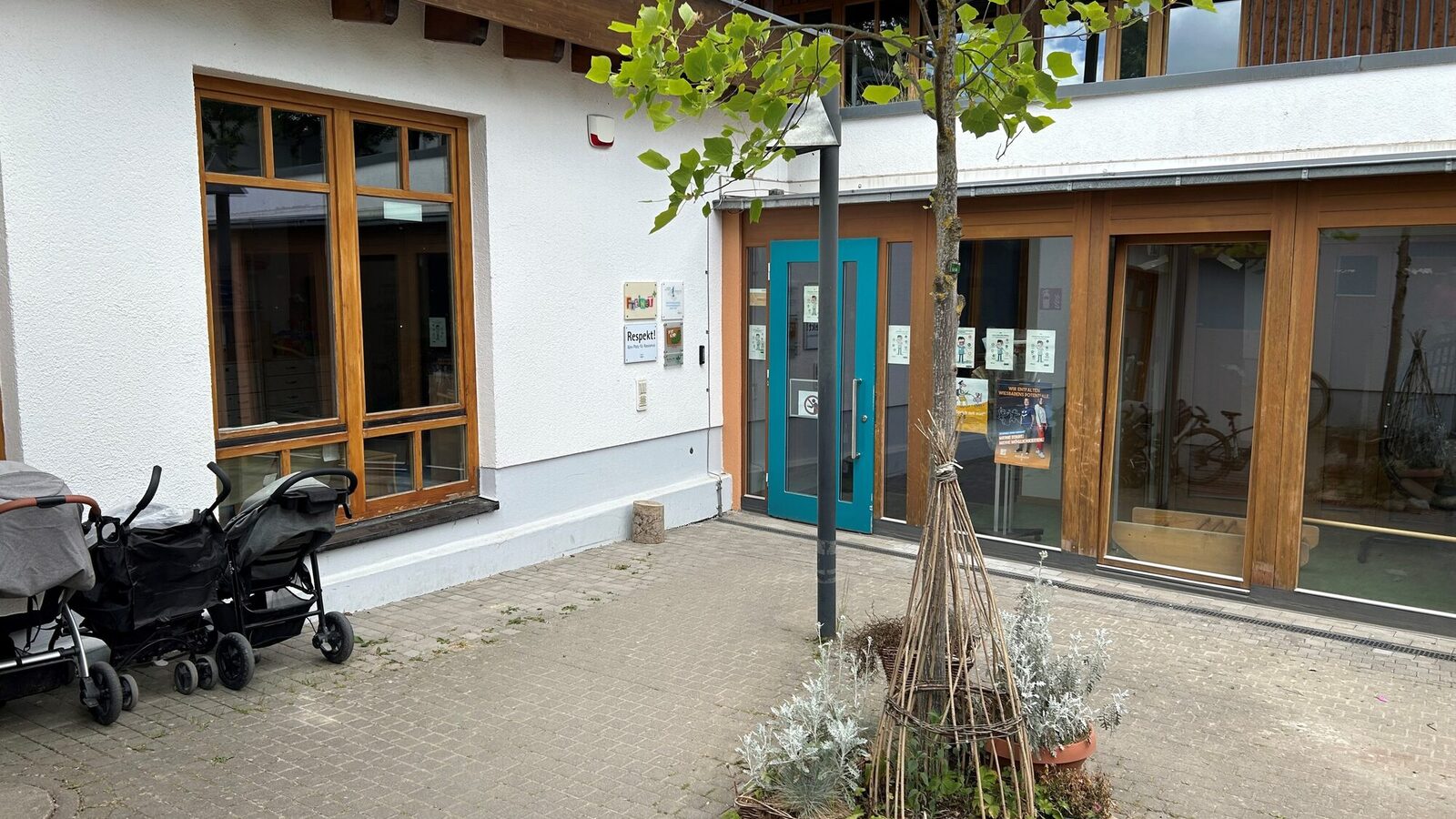 Der Eingang der Kindertagesstätte mit einem Baum im Vordergrund. Auf der linken Bildseite stehen Kinderwagen