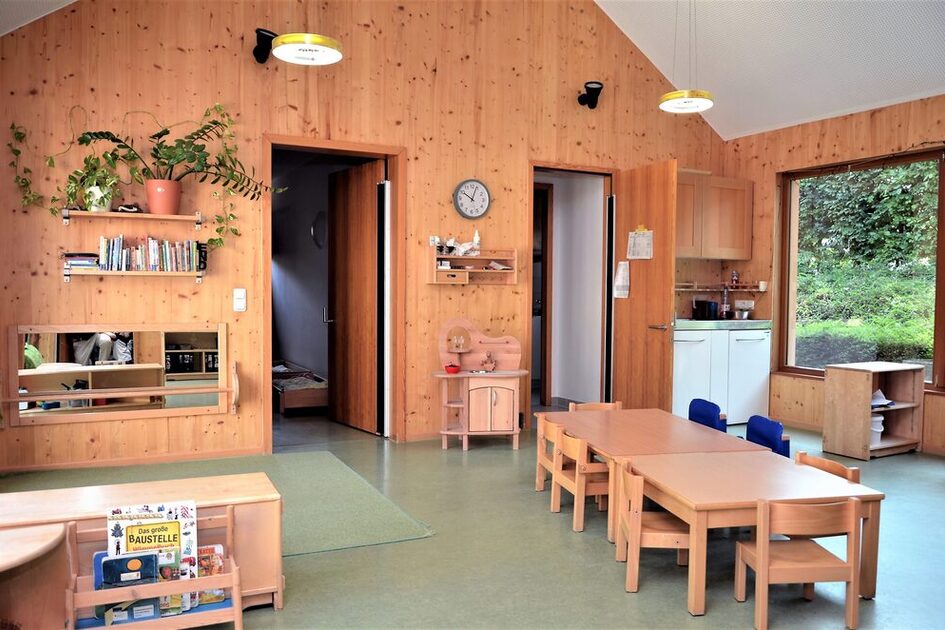 Ein Gruppenraum der Kindertagesstätte mit Tischen und Stühlen, einer kleinen Teeküche und einem Teppich auf dem Fußboden