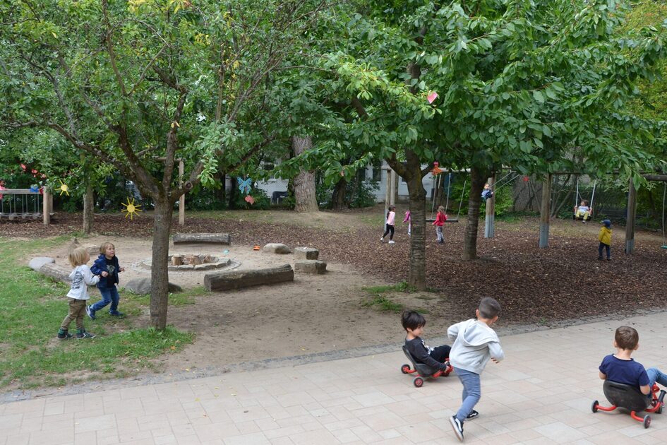 das Außengelände der Kindertagesstätte mit großen Bäumen und spielenden Kindern. Im der Bildmitte eine Feuerstelle