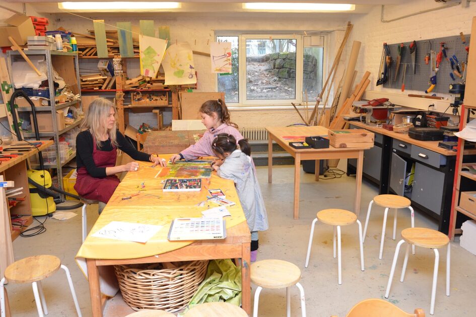 Die Werkstatt mit einem großen Arbeitstisch in der Mitte. An den Seiten stehen Werkbänke. Eine Erzieherin arbeitet mit zwei Kindern an dem großen Tisch