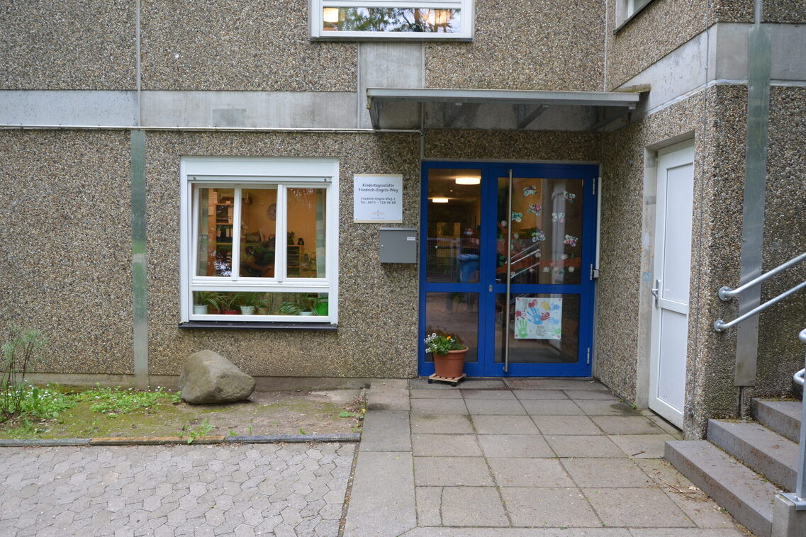 Der Eingangsbereich der Kindertagesstätte. Durch eine blaue Tür gelangt man in das Gebäude. Rechts führt eine Treppe zu dem oberen Teil des Außengeländes