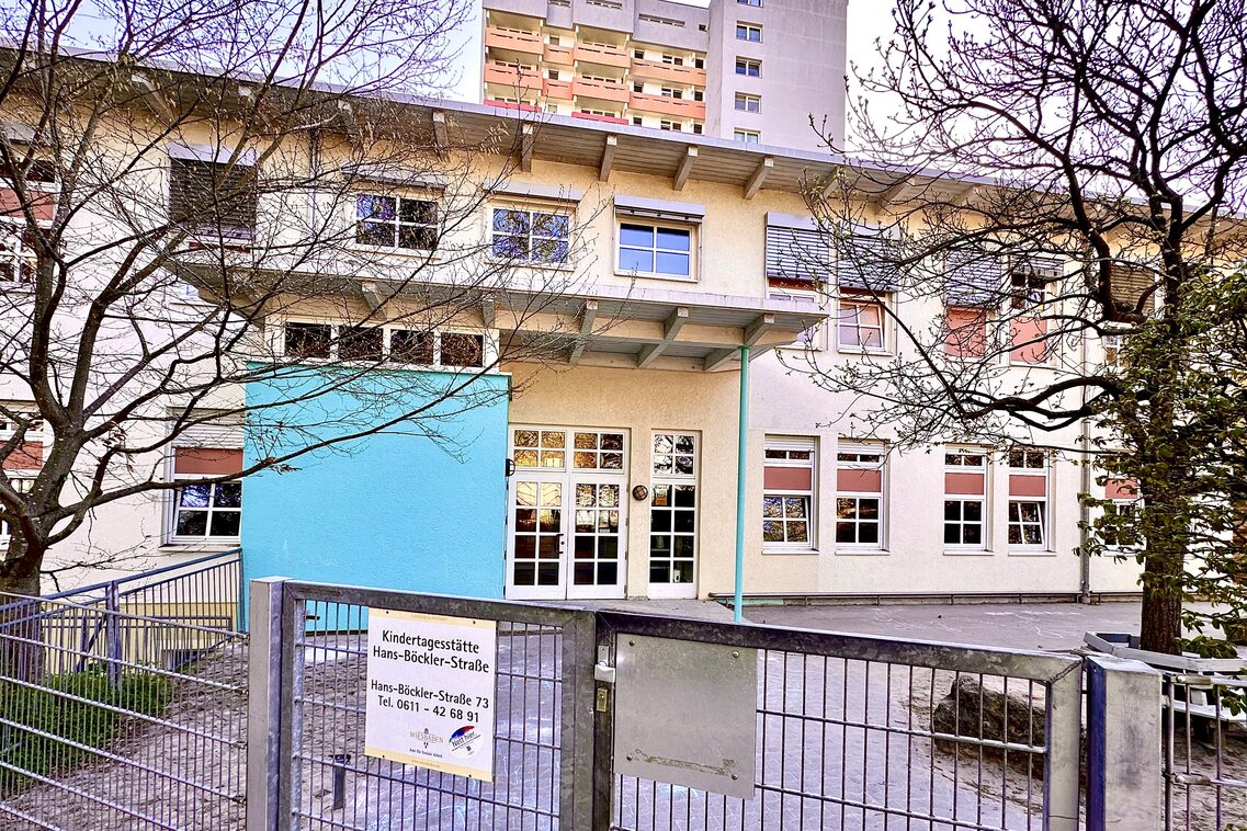 Der Eingangsberich der Kindertagesstätte. Am Zaun hängt das Schild mit dem Namen der Kita. das Gebäude mit einem überdachten Eingang steht im Hintergrund