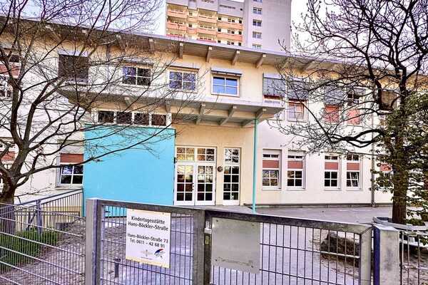 Der Eingangsberich der Kindertagesstätte. Am Zaun hängt das Schild mit dem Namen der Kita. das Gebäude mit einem überdachten Eingang steht im Hintergrund