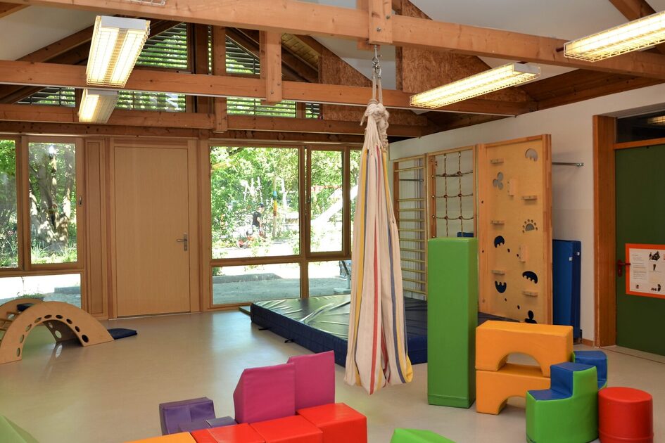 Der Bewegungsraum mit vielen mobilen Spielblöcken und einer Kletterwand. Eine große Fensterfront zeigt zum Außengelände