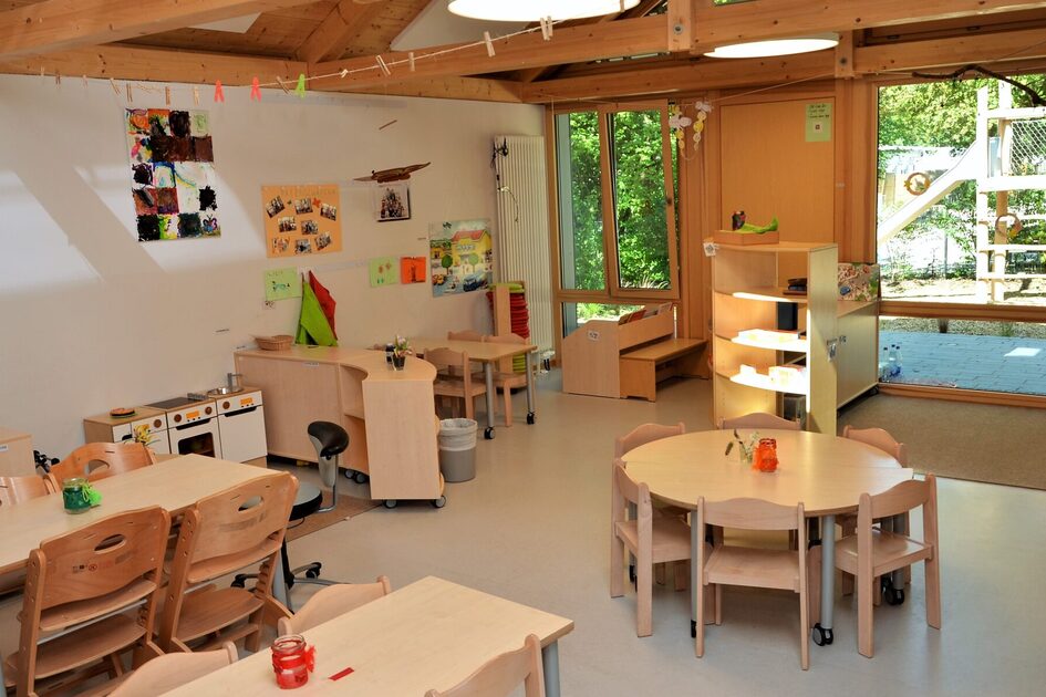 Ein Gruppenraum im Elementarbereich mit Tischen und Stühlen. An der Wand steht eine Spielküche. Ein großes Fenster zeigt zum Außengelände