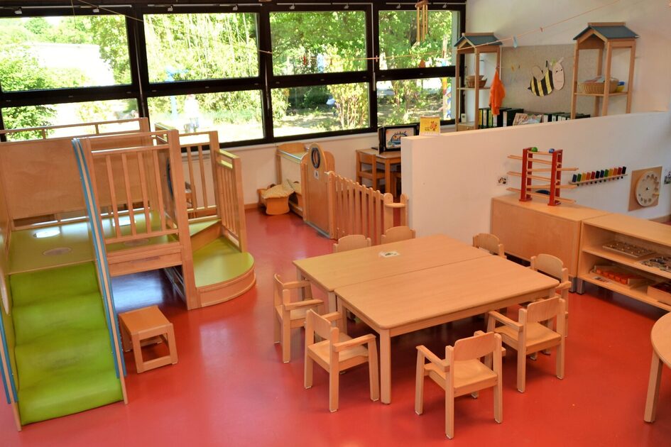 Eine Gruppe in der Krippe. In der Bildmitte steht ein niedriger Tisch mit kleinen Stühlen. Links ist ein kleines Kletterelement mit einer Rutsche