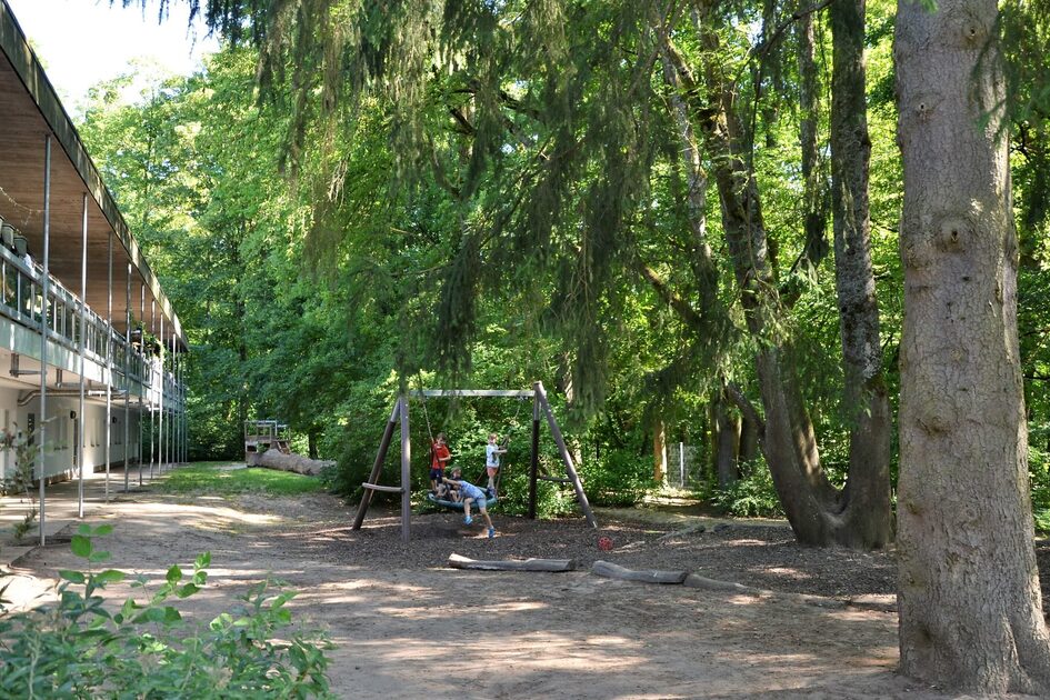 Das Außengelände der Kindertagesstätte. Links ist das Gebäude, rechts stehen große Bäume. In der Mitte ist eine große Nestschaukel mit Kinder.
