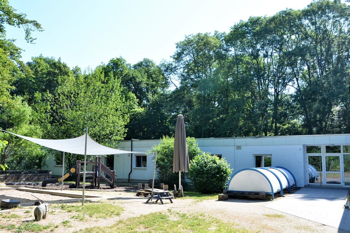 Der Garten der Kindertagesstätte mit dem Gebäude im Hintergrund. Links ist ein Spielbereich der mit einem Segel überdacht ist. Im Hintergrund stehen hohe Bäume