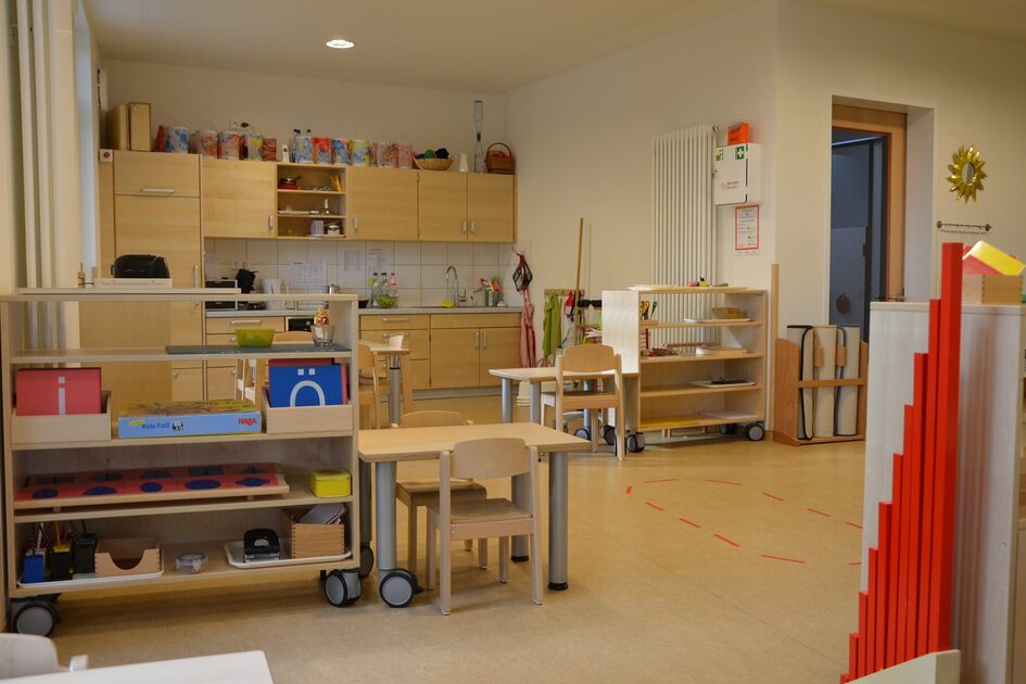 Eine Gruppe im Elementarbereich mit Tischen Stühlen und Regalen für Arbeitsmaterial. Im Hintergrund eine Küchenzeile.