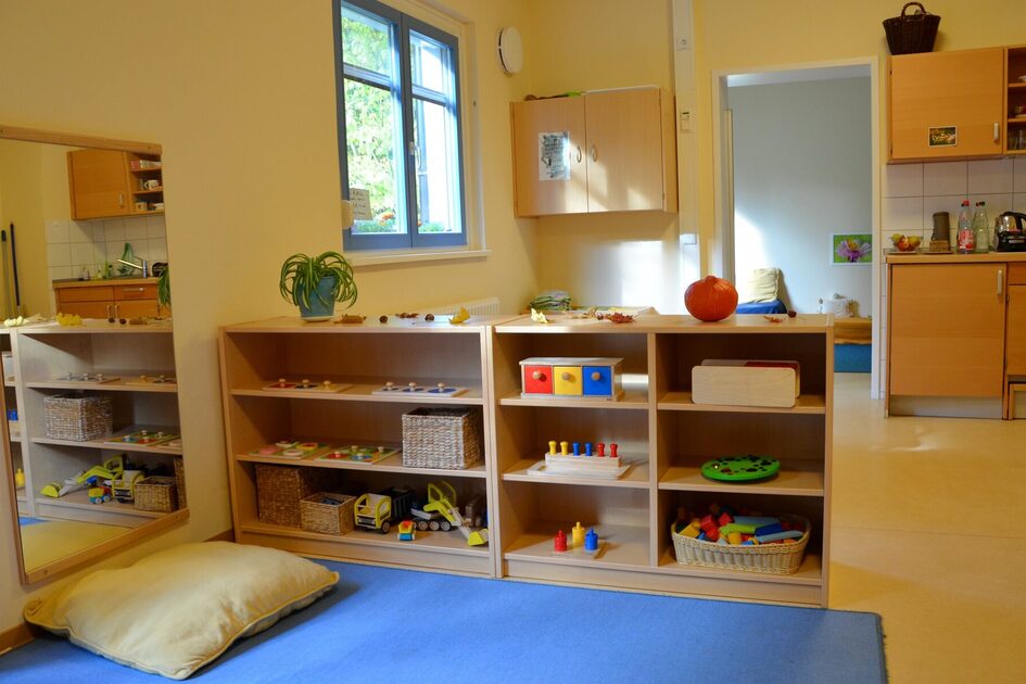 Eine Gruppe in der Krippe mit einem Regal für Spielmaterial. Davor liegt eine große blaue Matte. An der Wand hängt ein Spiegel.