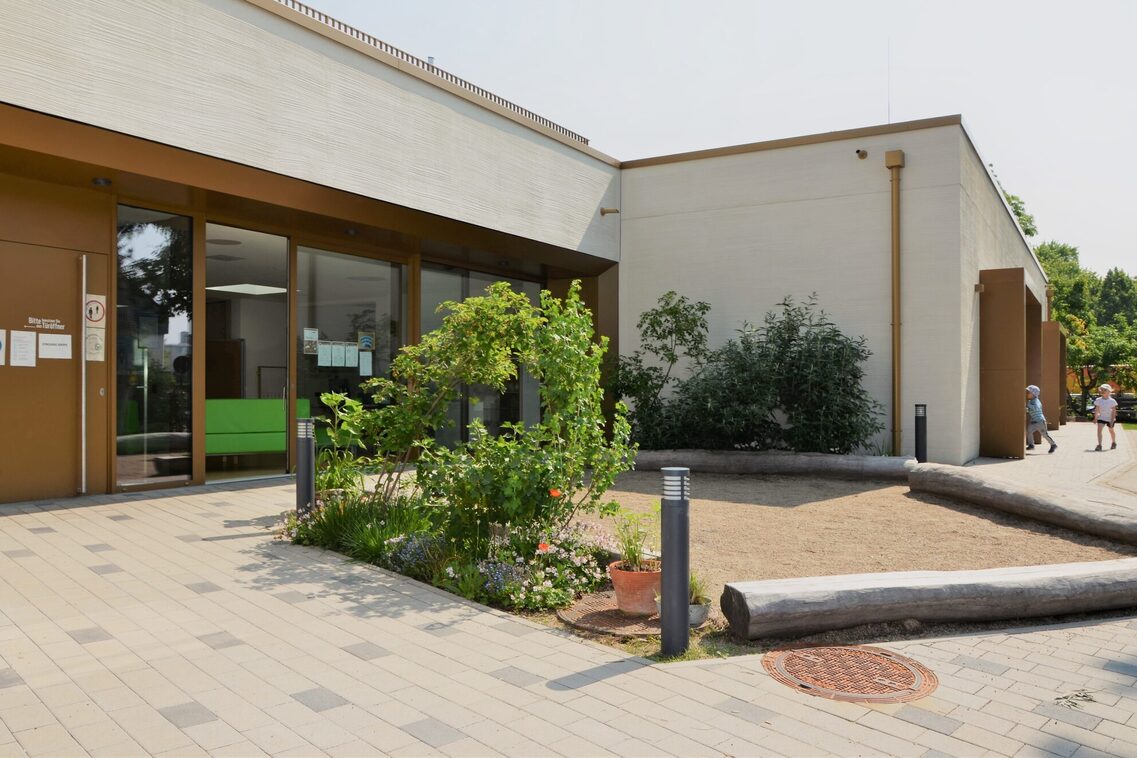 Der Eingangsbereich der Kindertagesstätte. Im Hintergrund das Gebäude mit einer großen Fensterfront, rechts stehen Büsche.