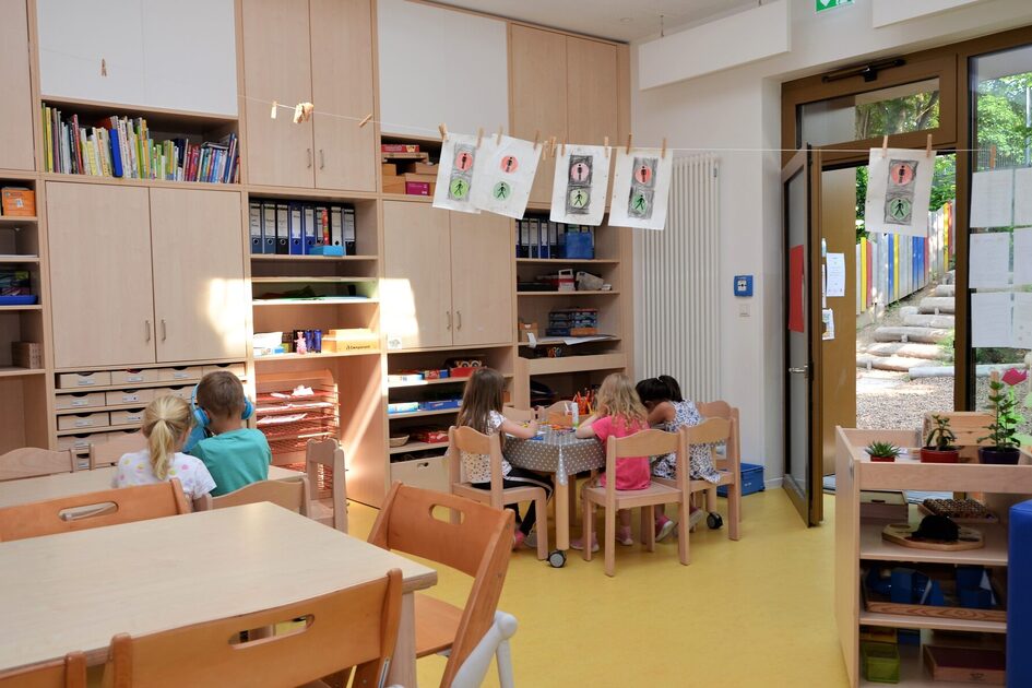 Ein Gruppenraum im Elementarbereich. An den Tischen sitzen Kinder und spielen oder malen. Im Hintergrund ein großer Einbauschrank mit Material