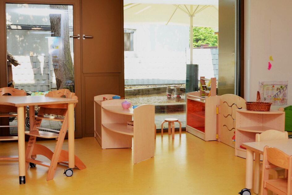Ein Gruppenraum in der Krippe. An einem Tisch stehen zwei Tripp-Trapp Stühle. Im Raum sind unterschiedliche Regale mit Elementen zum spielen.