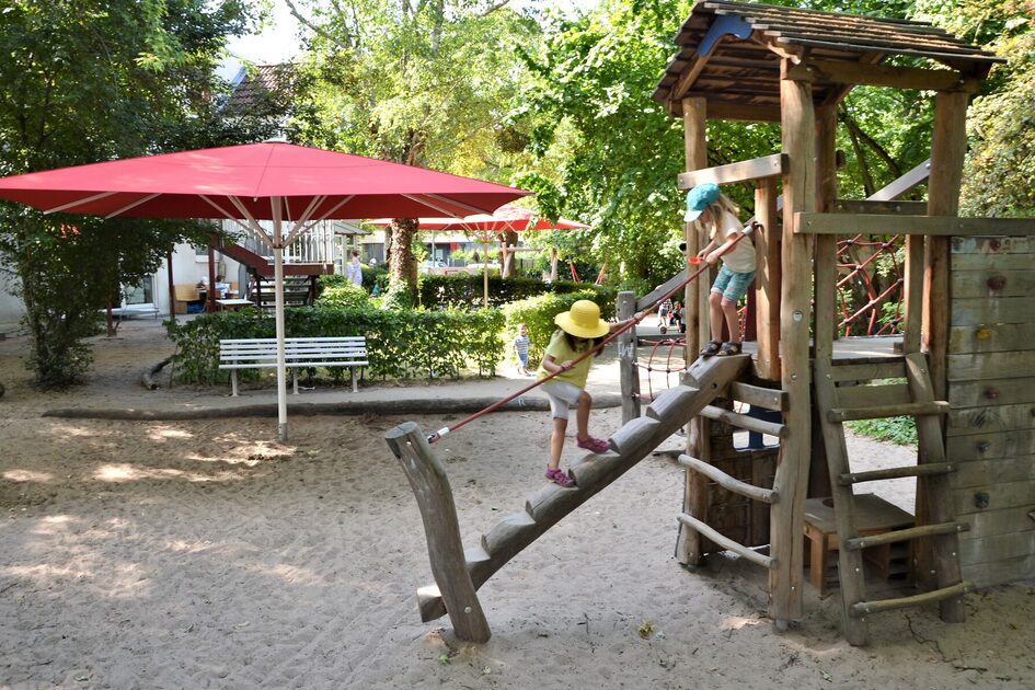 Das Außengelände mit dem Sandspielbereich und einem Kletterhaus mit zwei Kindern. Im Sand steht ein großer roter Sonnenschirm