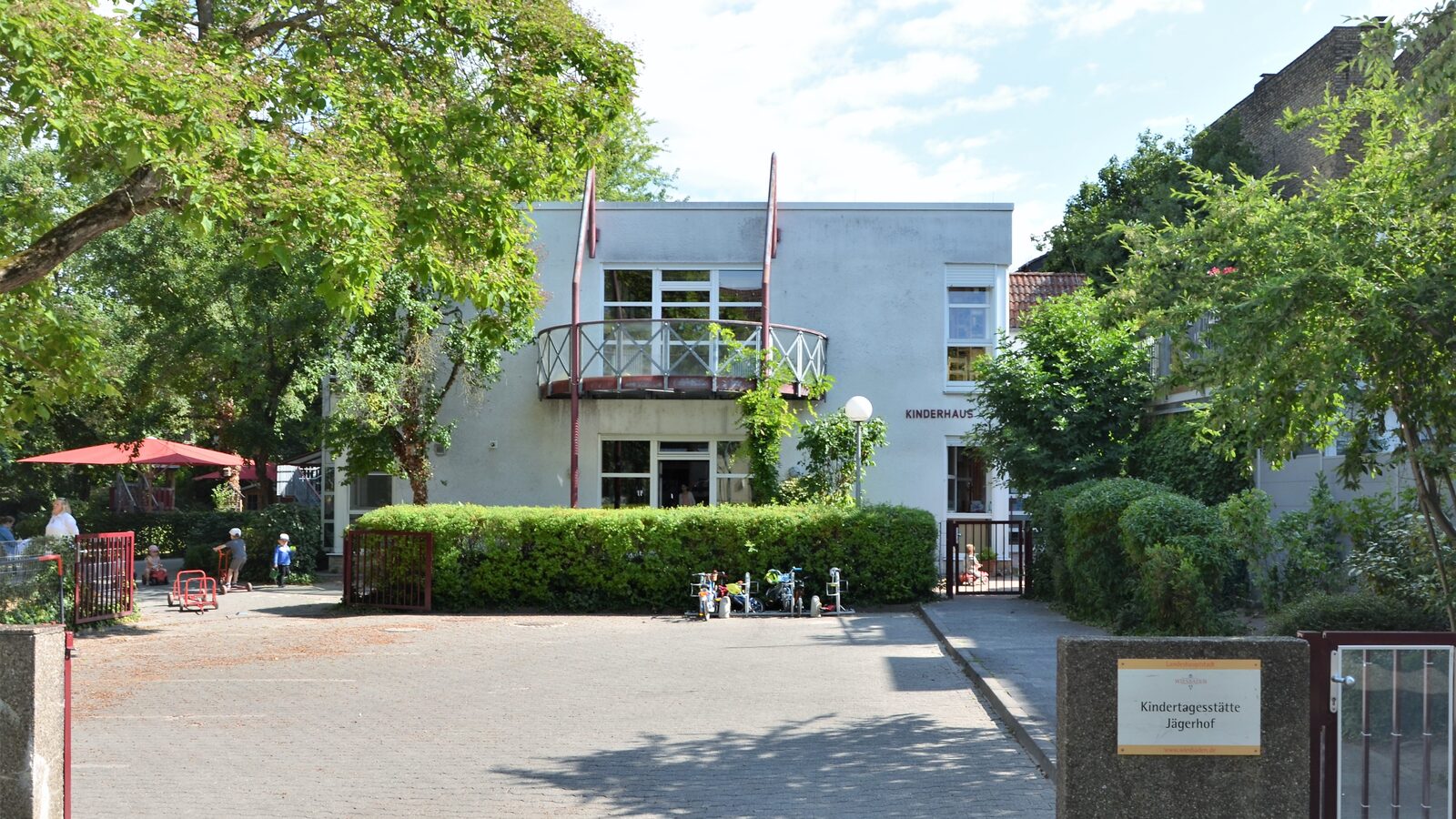 Der Eingang zur Kindertagesstätte mit einem kleine Hof. Im Hintergrund das Gebäude mit einer Hecke und einem Balkon im ersten Stock
