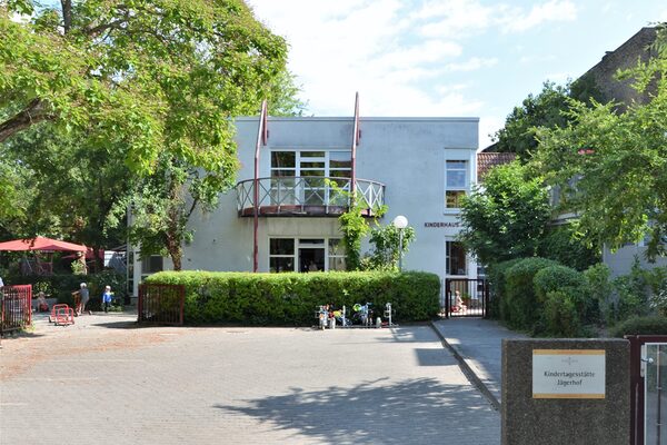 Der Eingang zur Kindertagesstätte mit einem kleine Hof. Im Hintergrund das Gebäude mit einer Hecke und einem Balkon im ersten Stock