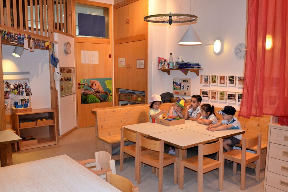 Ein gruppenraum der Kindertagesstätte. An einem Tisch sitzen fünf Kinder mit einem Spiel. In der linken Ecke sieht man den Beginn der zweiten Ebene