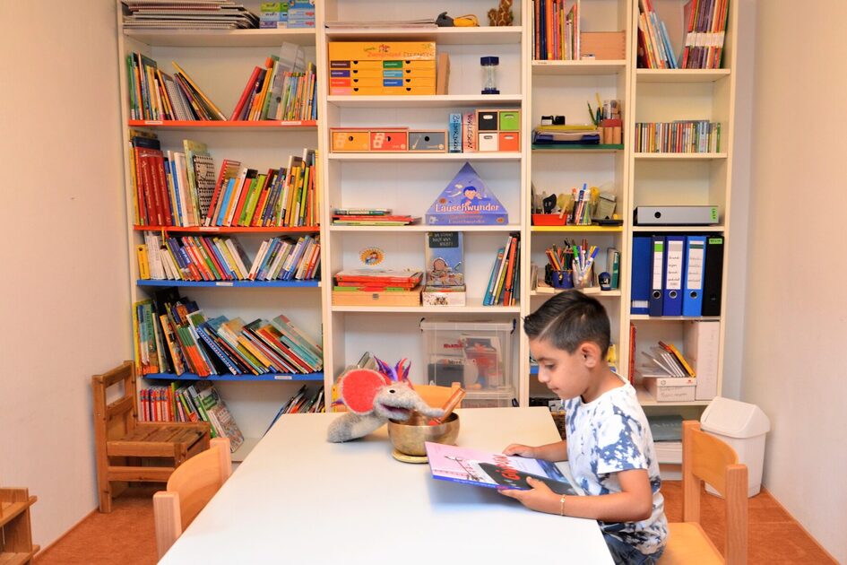 der Sprachraum der Kindertagesstätte. An einem Tisch sitz ein Junge und liest ein Buch. Im Hintergrund steht ein Bücherregal an der an der Wand