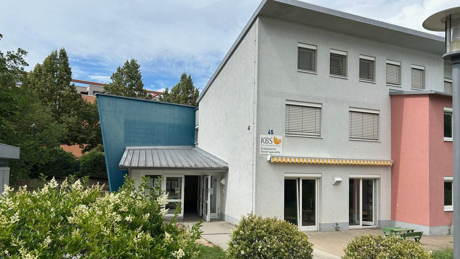 Eingangsbereich der Kindertagesstätte. Im Vordergrund stehen Büsche und an der Wand hängt ein Schild mit dem Logo der Kita