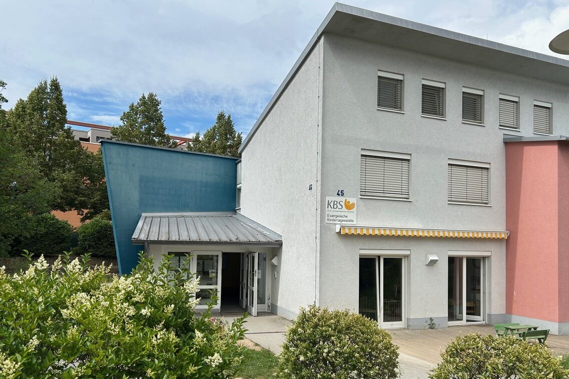 Eingangsbereich der Kindertagesstätte. Im Vordergrund stehen Büsche und an der Wand hängt ein Schild mit dem Logo der Kita