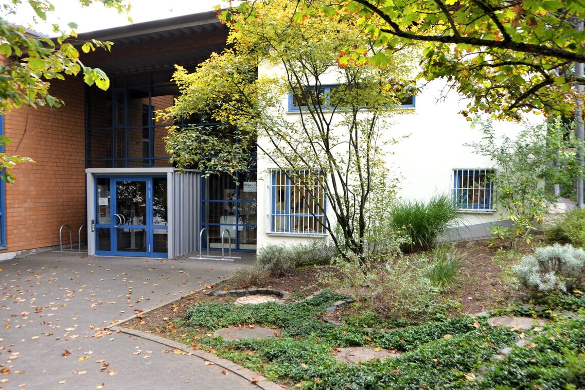 Der Weg zur Kindertagesstätte. Das Gebäude steht im Hintergrund. Auf der rechten Seite ein Blumenbeet mit einem Baum