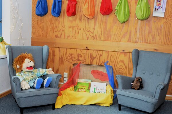 Die Bibliothek der Kindertagesstätte mit zwei kleinen Sessel und einem Lesekoffer. In einem Sessel sitz eine große Handpuppe