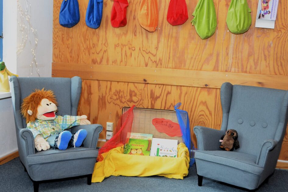 Die Bibliothek der Kindertagesstätte mit zwei kleinen Sessel und einem Lesekoffer. In einem Sessel sitz eine große Handpuppe